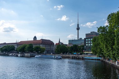 Almanya, 7 Haziran 2024. Büyük bir binası ve arka planında uzun bir kulesi olan bir şehir. Su sakin ve üzerinde yüzen tekneler var.