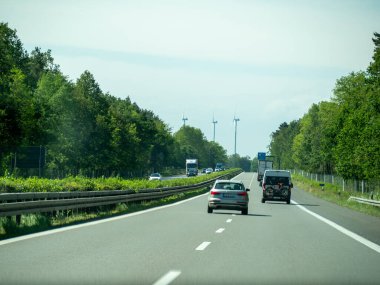 17 Mayıs 2024 'te arka planda bir sürü ağaç olan bir otoyolda bir araba ilerliyor. Araba yolun ortasındadır ve yolda başka arabalar ve kamyonlar da vardır. Gökyüzü açık.