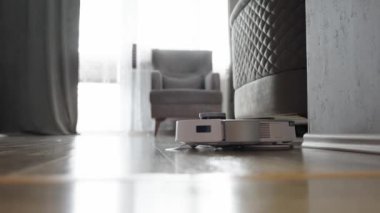 Elektrikli süpürge bir sandalyenin önünde yerde. Oda temiz ve düzenli, içeri ışık alan bir pencere var. Robot odanın ortasına yerleştirildi.