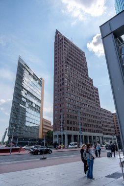 Bir grup insan yüksek bir binanın önündeki kaldırımda ayakta duruyor. Gökyüzü açık ve güneş parlıyor.