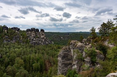 Arka planda bulutlu bir gökyüzü olan bir dağ sırası. Ağaçlar yemyeşil, dağlar sarp ve sarp.