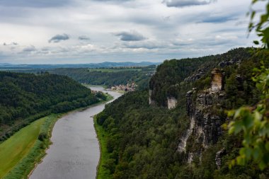 Arkasında kayalık bir uçurum olan yemyeşil bir vadiden bir nehir akar. Gökyüzü bulutlu, ortama dengesiz ve sakin bir atmosfer veriyor.