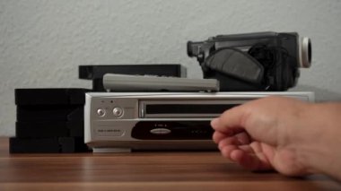 Küçük gümüş bir VHS oynatıcı bir yığın siyah kasetin yanında ahşap bir masada oturuyor. Gümüş ve siyah VCR.