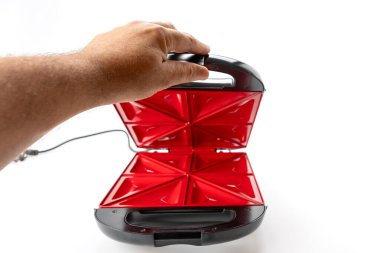 Bir kişi kırmızı ve siyah bir tost makinesi tutuyor. Tost makinesi açık ve kullanıma hazır.