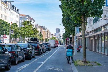 Almanya 7 Ağustos 2024. Mavi kulübeli bisikletli bir teslimatçı ile kalabalık bir cadde. Sokaklar park etmiş arabalar ve birkaç kamyonla dolu.