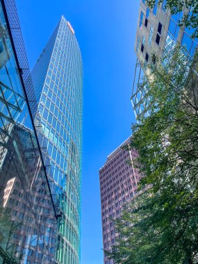 Aşağıdan cam cepheli yüksek bir bina görülüyor. Gökyüzü açık ve mavi ve ön plandaki ağaçlar yeşil. Modernlik ve entelektüellik kavramı, aynı zamanda açıklık hissi.