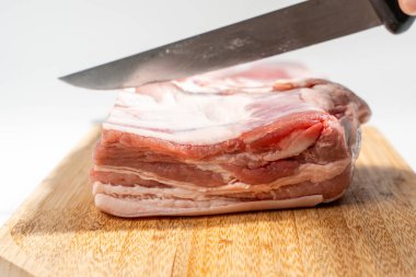 Bir insan ahşap bir kesme tahtasıyla bir parça et kesiyor. Bıçak etin üst kısmına yerleştirilmiş ve kişi onu hafif bir açıyla tutuyor. Beyaz arka planda izole edilmiş domuz eti..