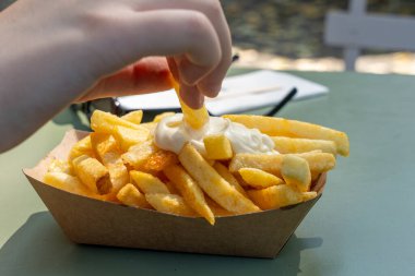 Birisi patates kızartmasını mayonez kabına daldırıyor. Patatesler altın sarısı ve çıtır çıtır görünüyor. Sahne rahat ve rahattır, basit bir aperatifin tadını çıkaran kişi...