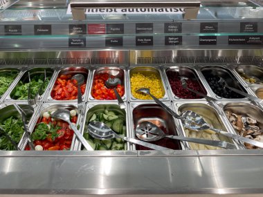 Farklı sebze ve meyvelerle dolu bir salata barı. Sebzeler küçük parçalara ayrılır ve farklı kaselerde düzenlenir. Kaseler gümüş bir tezgahın üzerine konur.