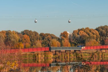 Nehrin üzerindeki yaya köprüsü. Köprü kırmızı ve suya yansıyor. Gökyüzü açık ve ağaçlar yapraklarla dolu.