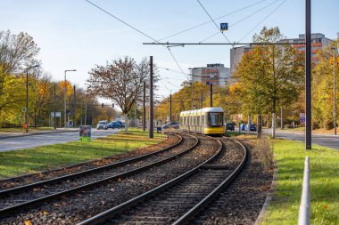 Almanya 3 Kasım 2024. Caddenin yanındaki raylarda bir tramvay hareket ediyor. Tramvay sarı ve beyazdır. Sokak ağaçlarla kaplı ve birkaç araba park halinde..