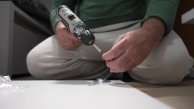 Elinde matkapla yerde oturan bir adam. Dolabı topluyor..