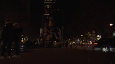 Almanya 17 Kasım 2024. Bir grup insan gece sokakta yürüyorlar. Sokaklar sokak lambalarıyla aydınlanıyor ve yan tarafa park edilmiş birkaç araba var. İnsanlar yürürken manzara sakin ve huzurlu.