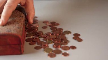 Bir adam ahşap bir sandığı açar, içine bir sürü bozuk para döker ve kapatır. Gri masada içinde bozuk paralar olan tahta bir sandık var..