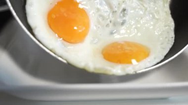 Üzerinde iki yumurta olan beyaz bir tabak. Biri diğerinden biraz daha büyük. Tabak masanın üstünde. Bir adam kızarmış yumurtaları tavadan tabağa aktarıyor..