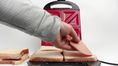 Sandviç yapmak için ekmeğe sosis koyar. Beyaz arkaplanda elektrikli sandviç makinesi.