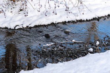 Karda bir su akışı yansıtılır. Su berrak ve sakin. Kar beyazdır ve zemini kaplar..