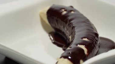 Üstü çikolata kaplı muz. Muz, çikolata kaplı bir tabakta, beyaz arka planda dönüyor..