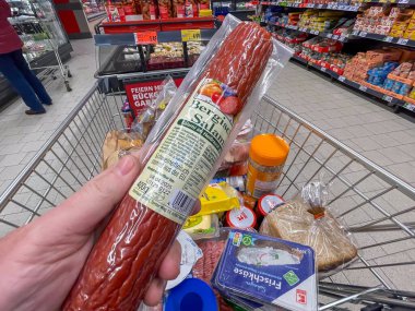 19 Şubat 2025 'te Almanya' da bir adam alışveriş arabasında sosis tutuyor. Sosis plastiğe sarılmış. Arabada bir kutu peynir ve bir şişe hardal da dahil olmak üzere başka eşyalar var.