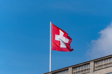 Üzerinde haç olan kırmızı ve beyaz bir bayrak gökyüzünde dalgalanıyor. Bayrak direğin üzerinde ve uzaktan görülebiliyor.