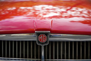 Almanya 11 Mayıs 2025. Kırmızı MG klasik radyatör ızgarası. Krom ızgara ve marka rozeti olan kırmızı bir MG klasik arabanın yakın çekimi. Kaput, ağaçları ve gökyüzünü sıcak tonlarda yansıtıyor..