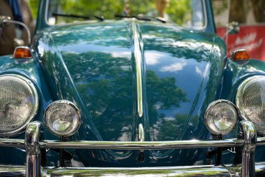 Almanya 11 Mayıs 2025. Mavi renkli VW Beetle 'ın önden görünüşü ve ikiz ışıkları. Mavi klasik bir VW Beetle 'ın simetrik farları ve yardımcı sis ışıklarını gösteren ikonik ön görüntüsü. Gökyüzünün ve kaputtaki ağaçların temiz yansımaları.