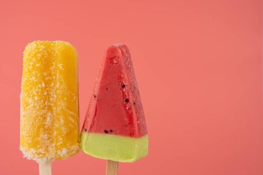 Pembe arka planda yan yana iki renkli meyve dondurması. Solda sarı dondurma, sağda yeşil kaplamalı karpuz dondurması. Kombinasyon eğlenceli, yazlık ve davetkar görünüyor..