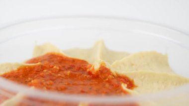 Kırmızı biber soslu humus temiz bir plastik kabın içine konur ve beyaz bir zemin üzerinde döner. Kompozisyon taze ve iştah açıcı görünüyor..