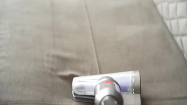 Açık gri bir kanepeyi temizliyorum. Hafif kanepe, el elektrikli süpürgesi ile yavaşça ve eşit bir şekilde temizlendi. Yumuşak madde yumuşak bir ışığı yansıtır..