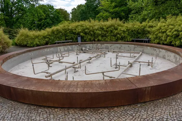 Paslı metal kenarlı boş, yuvarlak bir çeşme iyi korunmuş bir parkın ortasında duruyor. İçerideki su boruları görülebilir durumda, bu da bakım ya da kapatma anlamına geliyor..