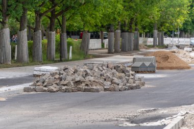 Asfalt ve kumdan yapılmış inşaat malzemeleri. Bir yığın kaldırım taşı, kaldırım taşları ve bir dağ dolusu kum yeşil bir caddedeki inşaat alanını işaret ediyor. Görüntü, kentsel yol yapımının ilerlemesini belgeliyor.