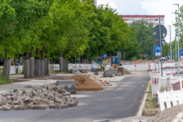 Yol İnşaat Alanı, Makine ve Bariyerler. İçinde kazıcılar, tabelalar ve inşaat malzemeleri olan bir inşaat alanı. Arka planda yeşil ağaçlarla çevrili bir yerleşim yeri var..