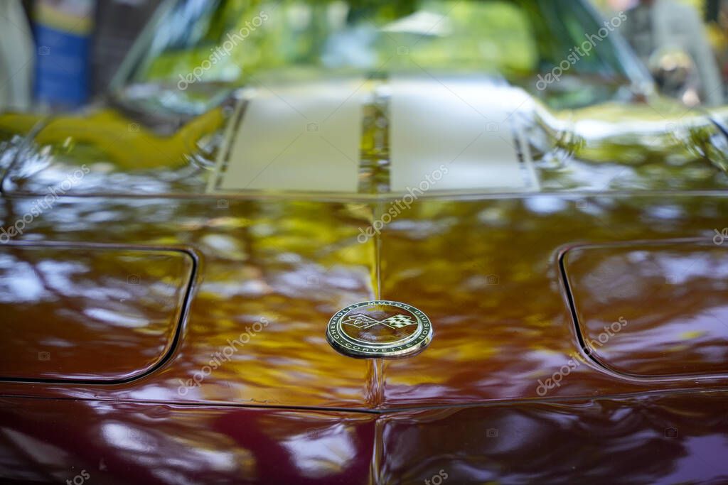 Corvette hood with emblem. The hood gleams in deep red and bears the signature Corvette emblem with crossed flags. Reflections of the surroundings dance on the paint.