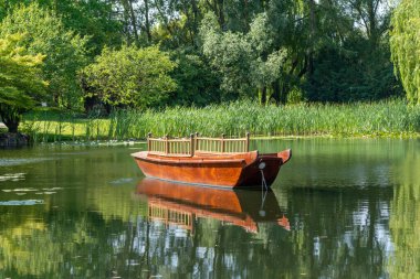 Kırsal bir ahşap tekne ayna gibi pürüzsüz bir gölette huzur içinde yüzer. Yeşil kıyı bitkileri ve ağaçlar suya yansıyor..