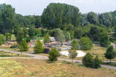 Yeşillik ve yürüyüş yollarıyla çevrili, çocuklarla dolu modern bir oyun alanı. Arka planda ağaçlar ve şehir binaları var..