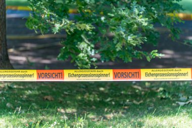 Meşe işlemeli güve uyarı musluğu. Bir uyarı bandı meşe güvelerinin oluşturduğu tehlikeyi gösteriyor. Çevrede meşe ağaçları ve güneşli bir park manzarası var..