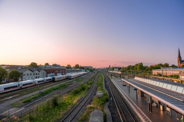 Gündoğumunda modern ve tarihi trenlerle genişleyen bir tren istasyonuna bakın. Arka planda yeşil şehrin üzerinde bir kilise kulesi var..