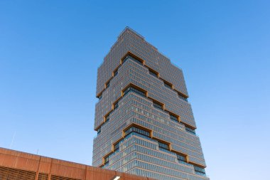 Modern, üst üste yığılmış bir kule mavi gökyüzüne yükseliyor. Ön cephe, altın renkli elementlerle simetrik cam yüzeylerden oluşuyor..