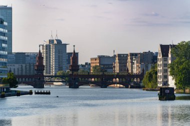 Berlin 'deki Oberbaum Köprüsü' nde sarı bir metro treni var. Arka planda, Spree kıyısındaki modern ve tarihi binalar.