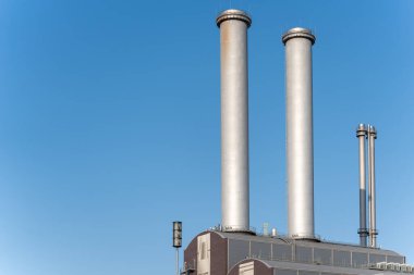 Mavi gökyüzüne doğru yükselen iki büyük sanayi bacası. Modern tesis, dikey mimarisiyle şehir manzarasına hakim..