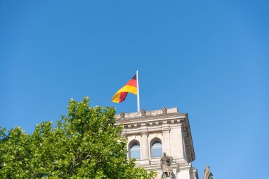 Alman ulusal bayrağı heykel ve sütunlarıyla tarihi bir binanın tepesinde dalgalanıyor. Parlak mavi gökyüzü ve yeşil ağaç yaprakları sahne çerçevesi.