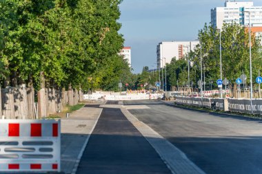 Yeni asfalt bir sokak, bariyerlerle kapatılmış bir inşaat alanına götürür. Yeşil ağaçlar caddede ve yüksek binalar uzaktan görülebiliyor..