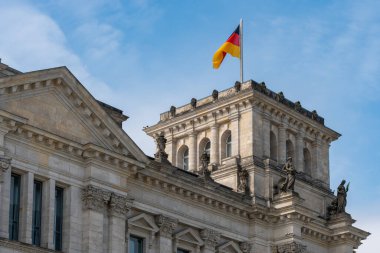 Mavi gökyüzünde dalgalanan bayrağıyla Alman Bundestag 'ı. Binanın üzerindeki heykeller tarihsel önemini vurguluyor..
