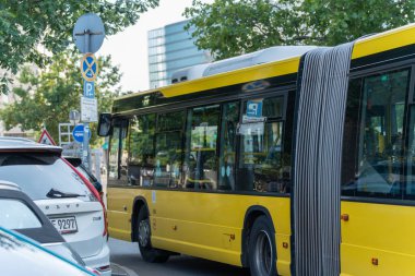 Almanya 19 Temmuz 2025. Sarı eklemli bir BVG otobüsü kalabalık bir şehir ortamında park halindeki arabaların ve Alman sokak tabelasının yanından geçiyor. Modern trafik ve toplu taşıma araçlarının bir karışımı yeşil ağaçların altında görülebilir.