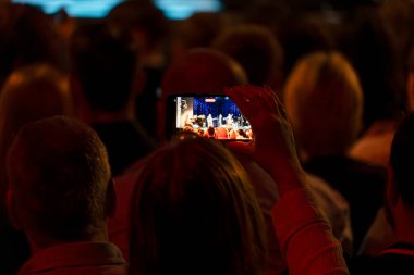 Kırmızı ışıklandırmalı loş bir ayarda, birisi bir sahne şovunu akıllı telefondan yakalıyor. Ekranda canlı bir hoparlör grubu ya da grubu gösteriliyor.
