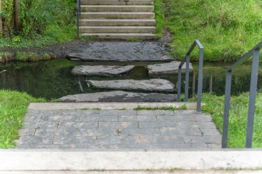 Akıntıdaki Basamaklar. Taş levhalardan yapılmış dar bir patika sığ bir dereye çıkıyor. Sahne sakin görünür ve duraksamayı davet eder.