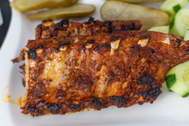 Izgara kaburga açılı manzara. Kömürleşmiş kabuklu ızgara pirzola ve sarımsak parçaları turşu ve taze salatalık dilimlerinin yanında beyaz bir tabakta dinleniyor. Yumuşak ve lezzetli et, tadını çıkarmaya hazır..