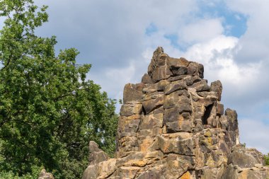Bulutlu gökyüzü ile doğal kaya oluşumu. Büyük yıpranmış kaya oluşumu yeşil ağaçlardan oluşan bir zemin ve parçalı bulutlu bir gökyüzüne doğru yükseliyor. Engebeli dokusu eski vahşi doğayı çağrıştırıyor..