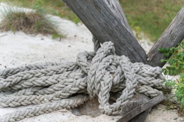 Kalın, örgülü bir ip kumlu zeminde, ahşap bir direğin etrafına sarılmış. Sahne, bir deniz manzarasını anımsatıyor..