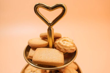 Danimarka tereyağlı kurabiyelerin yakın çekimi altın servis tepsisinin en üst katında, kalp saplı. Şeftali arka planı sıcak ve davetkar bir atmosfer yaratıyor..